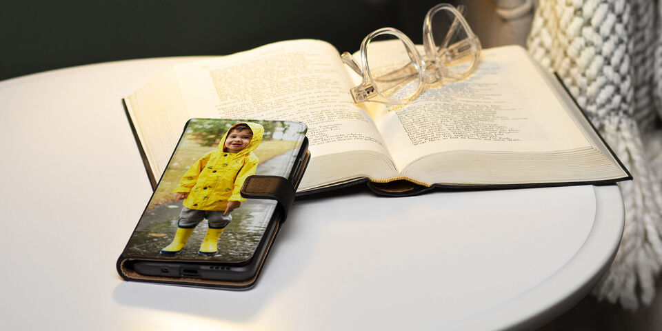 Un smartphone sur une table montre un enfant joyeux dans un manteau de pluie et des bottes. À côté du téléphone, un livre ouvert et des lunettes.
