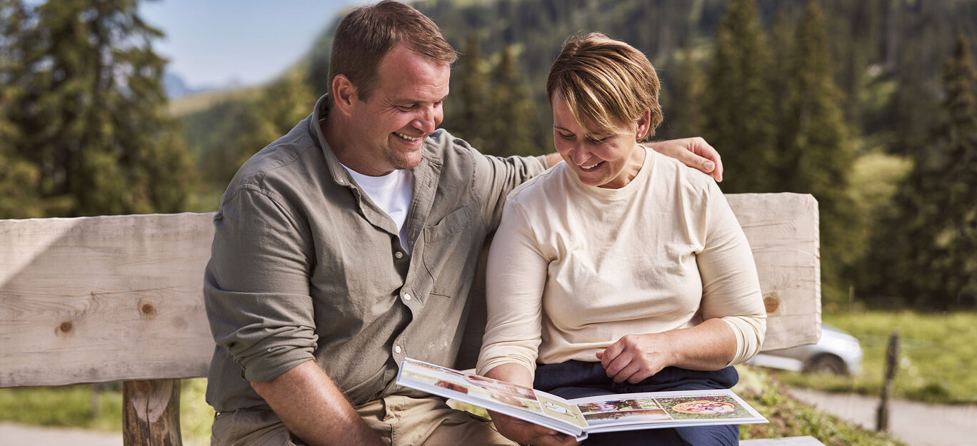 Un couple examine ensemble un album photo assis sur un banc dans la nature.