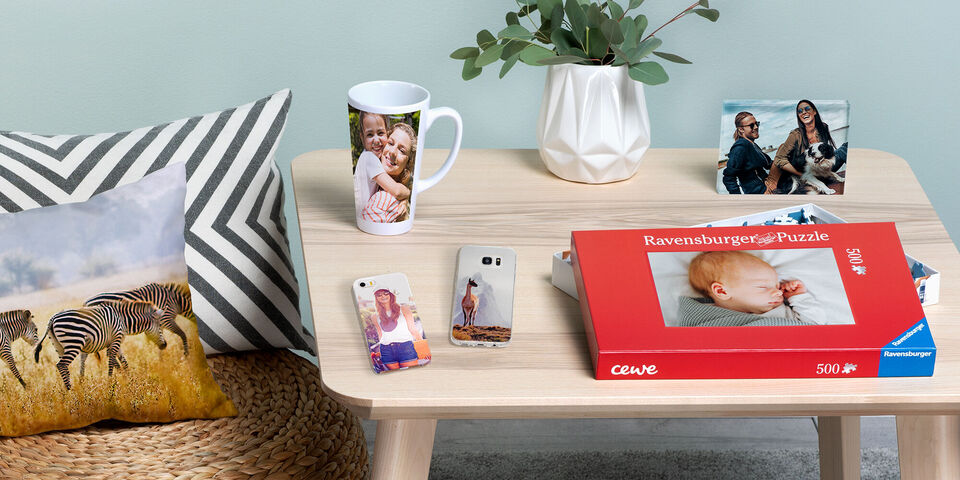 Une table avec divers produits photo : un livre photo CEWE, une tasse avec une photo, des coques de téléphone et un puzzle. Un coussin avec motif zèbre et des plantes complètent la scène.