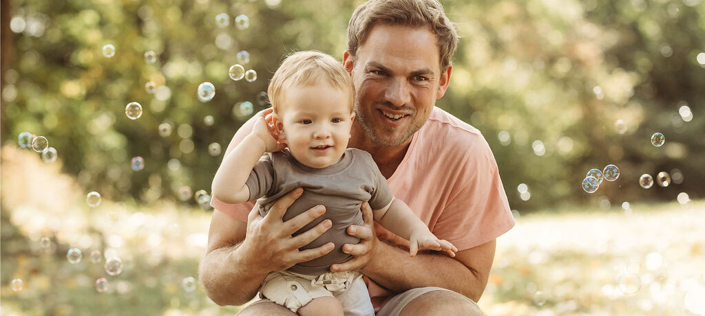Un père tient son petit enfant dans les bras, tous deux sourient joyeusement à la caméra. Des bulles colorées les entourent.