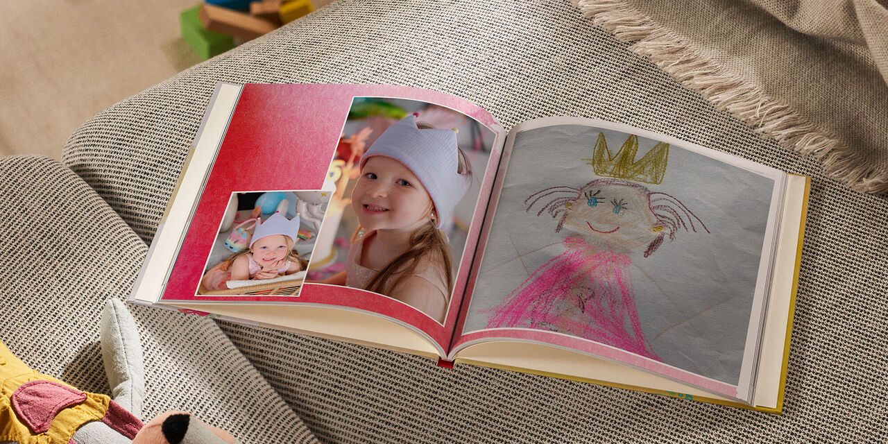 Sur un sofa, un LIVRE PHOTO CEWE ouvert est posé à côté d’un animal en peluche et d’une couverture. La page de gauche montre deux photos d’une fillette souriante portant une couronne. Sur la page de droite, on peut voir un dessin d’enfant représentant une princesse coiffée d’une couronne. L’arrière-plan de la double page est décoré d’un motif à l’aquarelle rouge.